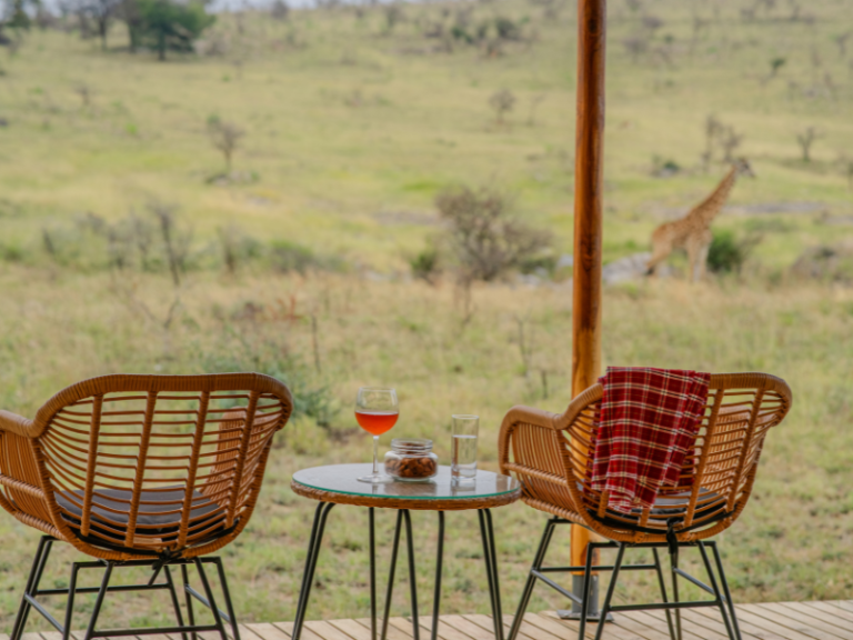 Two wicker chairs and a small table set with drinks on a safari deck, overlooking open plains where a giraffe wanders in the distance.