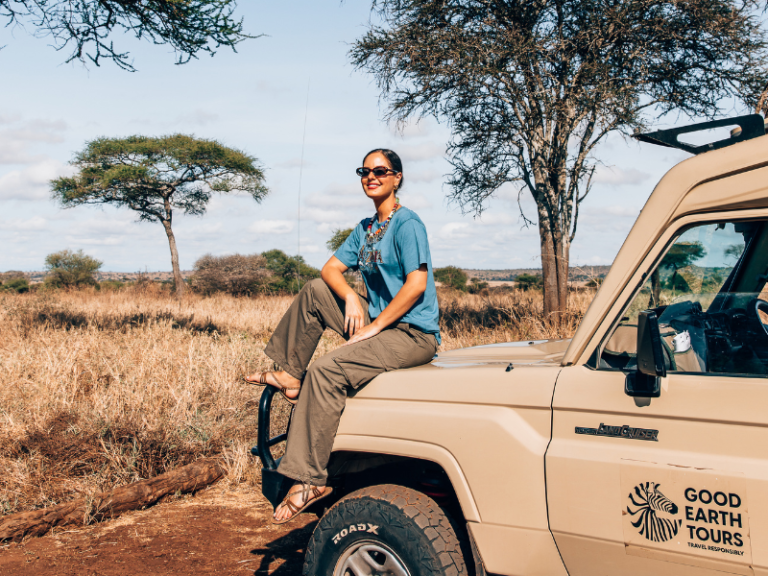 Traveler sitting on the hood of a safari vehicle with Good Earth Tours branding, enjoying the open savannah scenery.