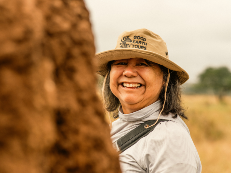Guest smiling warmly beside a termite mound while wearing a safari hat on a sunny day in the bush.
