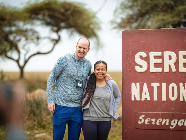 Smiling couple posing beside the Serengeti National Park entrance sign on a sunny safari day.