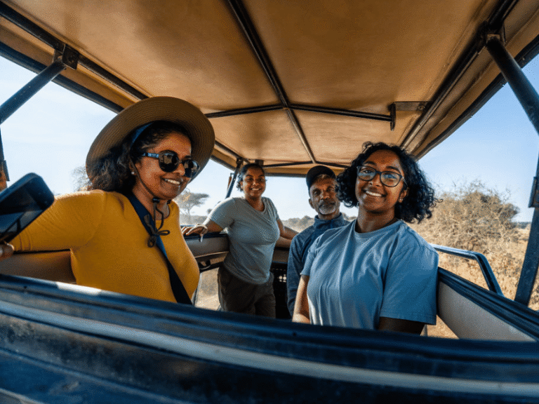 A smiling group of travelers enjoying their Tanzania safari inside an open-roof safari vehicle, with golden savanna in the background.