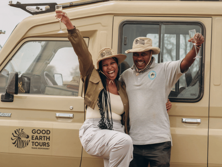 Two safari guests celebrating with champagne next to a Good Earth Tours vehicle in Tanzania, wearing safari hats and big smiles.