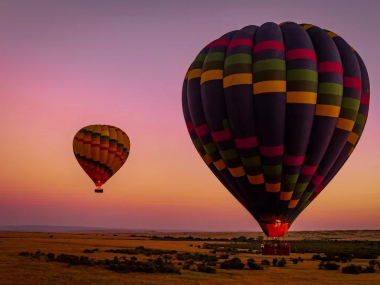 Colorful hot air balloons rising over the savannah at dawn, glowing against a pink and purple sky.