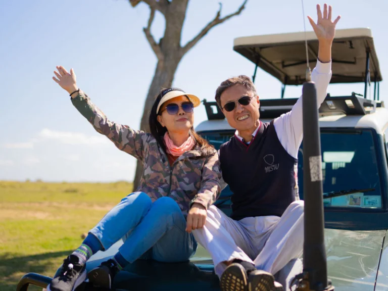 Couple waving joyfully while sitting on the hood of a safari vehicle in wide open grasslands.