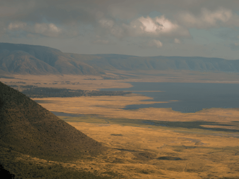 Sweeping view of Ngorongoro Crater in Tanzania with grasslands, water, and distant hills under a partly cloudy sky.