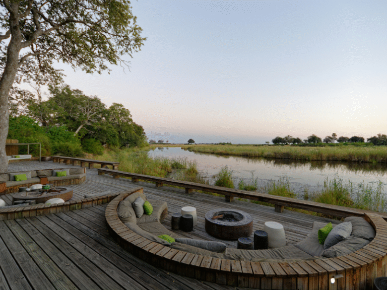 Outdoor seating area of a luxury safari lodge overlooking a calm river in Botswana at dusk.