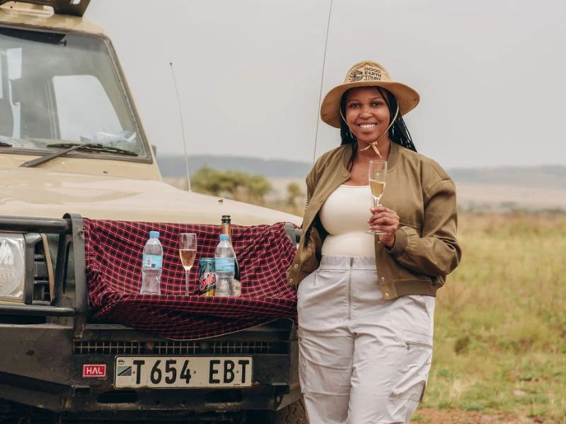 Safari Khaki Clothes: Do You Really Need Neutral Colors on Safari? 2 girl smiling & holding a champagne flute infront of a safari vehicle, wearing safari clothing