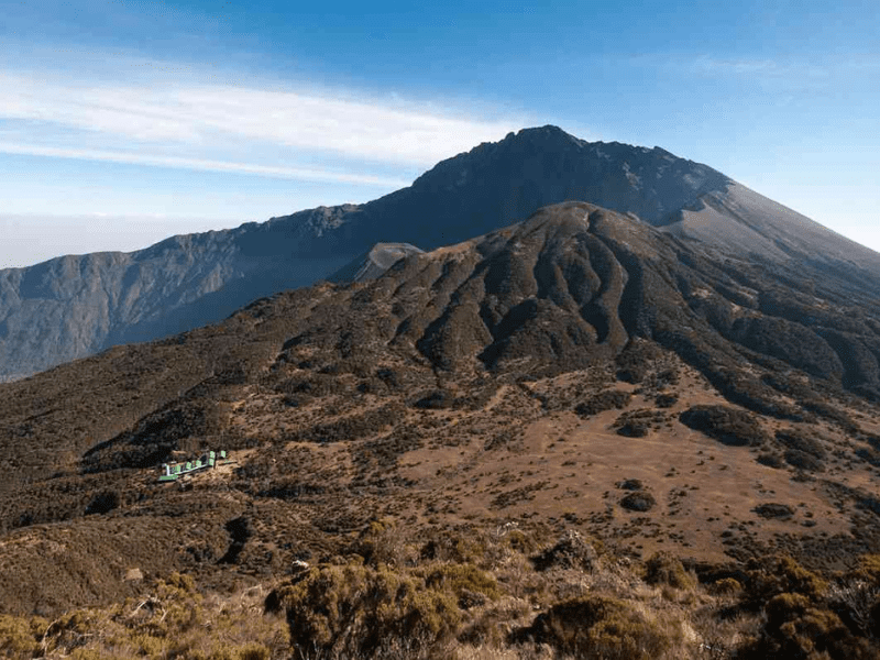 Scenic view of Mt Meru with alpine moorland in the foreground and a distant hut visible on the Saddle Hut route, a popular trail for climbing Mt Meru Tanzania.