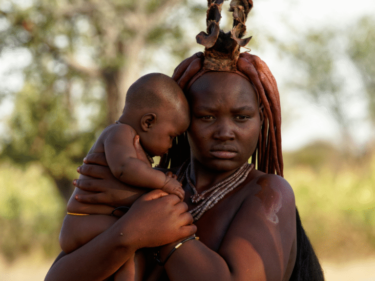 Himba woman holding her baby outdoors, wearing traditional ochre adornments and jewelry in northern Namibia.
