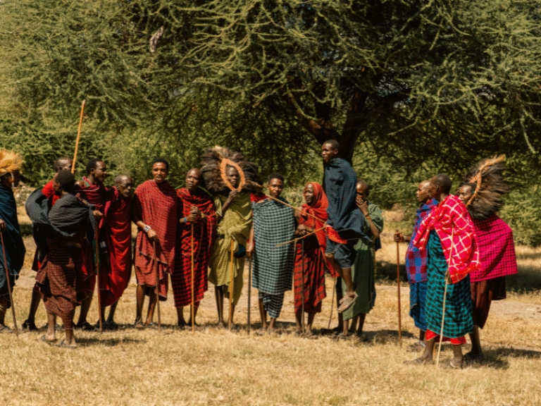 A group of Maasai men in colorful shuka cloths performing a traditional jumping dance under the shade of an acacia tree in northern Tanzania.