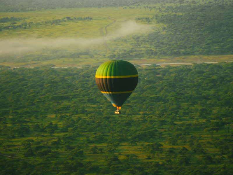 A green and yellow hot air balloon safari tour floats above the lush plains of Tanzania at sunrise.
