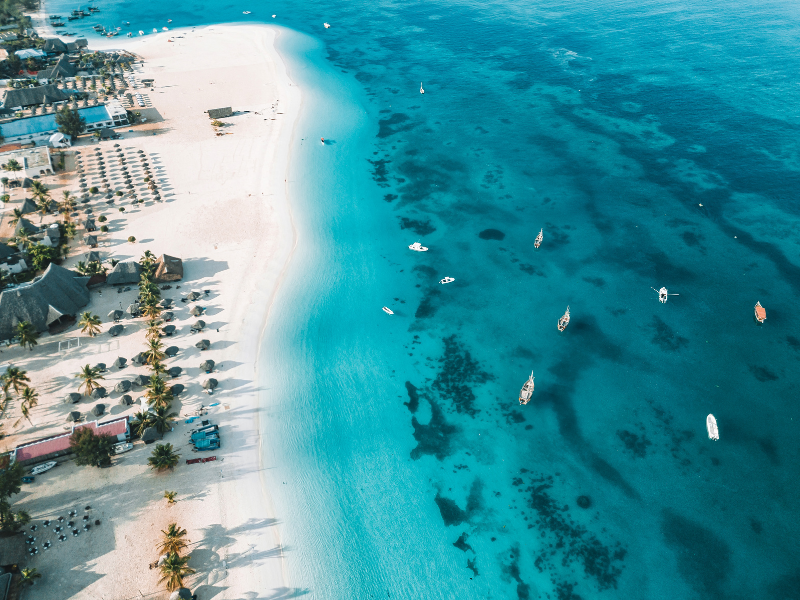 Aerial view of a pristine white sand beach in Zanzibar, Tanzania, with turquoise ocean waters and scattered boats offshore.
