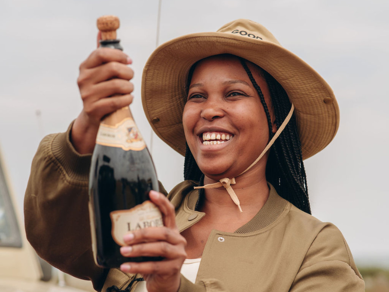 What’s Included in Luxury Tanzania Safaris (and Why They’re Worth It) 3 Smiling safari guide in a wide-brimmed hat holding a bottle of sparkling wine during a celebratory bush moment.