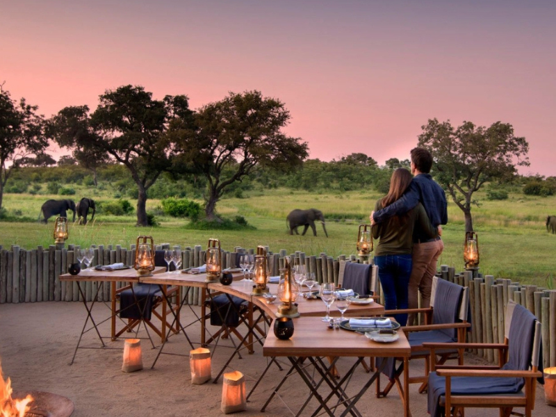 What’s Included in Luxury Tanzania Safaris (and Why They’re Worth It) 1 Couple standing arm in arm beside a lantern-lit bush dinner setup as elephants wander across the grassy plains at dusk, capturing the romance of luxury Tanzania safaris.