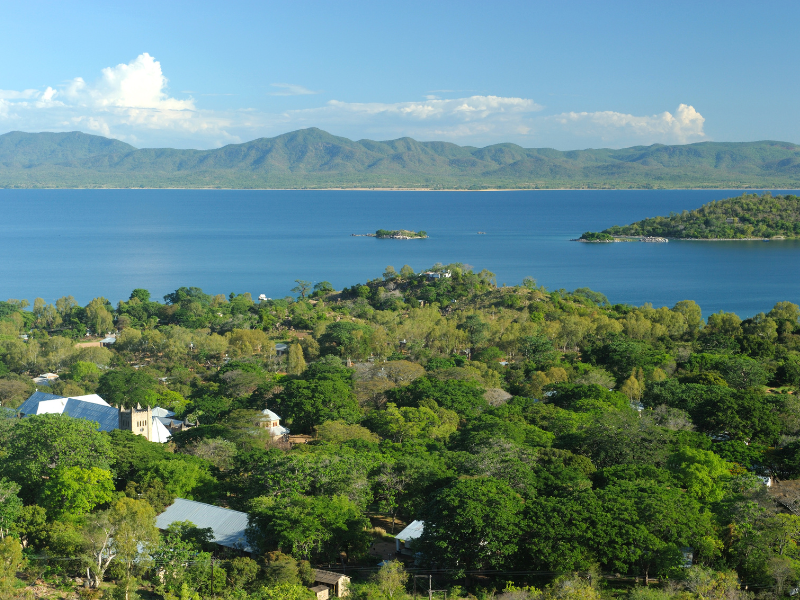 Aerial view of Lake Malawi’s shoreline near Mozambique, with lush green trees, scattered homes, and mountains in the background.