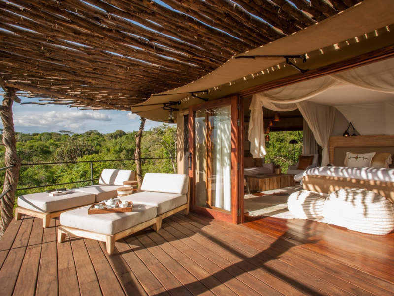 What’s Included in Luxury Tanzania Safaris (and Why They’re Worth It) 4 Open-air safari suite with a shaded wooden deck, cozy loungers and a canopy bed, surrounded by lush green treetops.