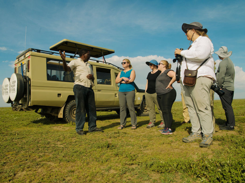 Safari guide speaking to a small group of travelers beside a pop-top Land Cruiser on open grassland.