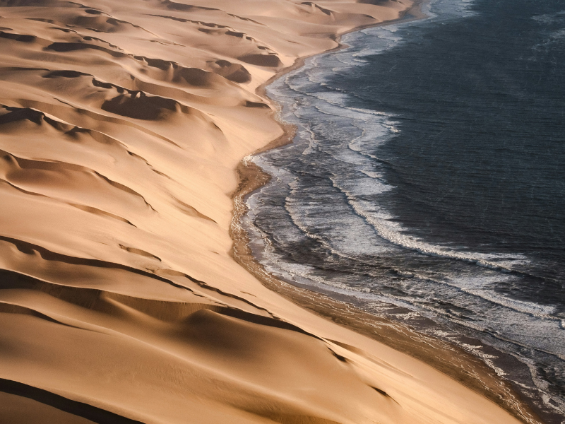 Aerial view of golden sand dunes meeting the Atlantic Ocean along Namibia’s Skeleton Coast, where desert landscapes blend with the sea.