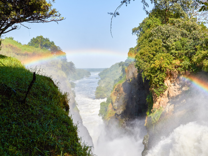 A stunning view of Murchison Falls National Park in Uganda, where the Nile River crashes through a narrow gorge creating a powerful waterfall and misty rainbow.