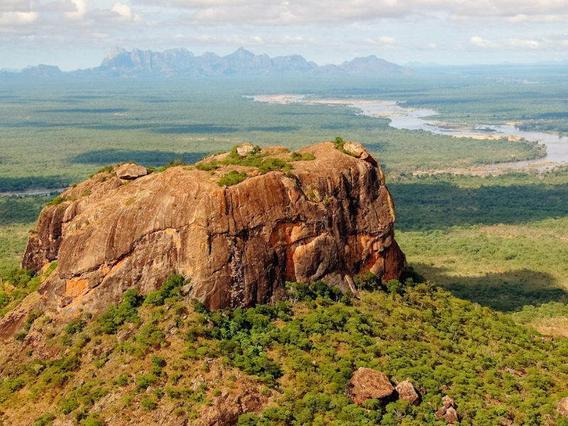 Aerial view of Niassa Reserve with green bushland, rocky outcrops and winding riverbeds under a bright sky.