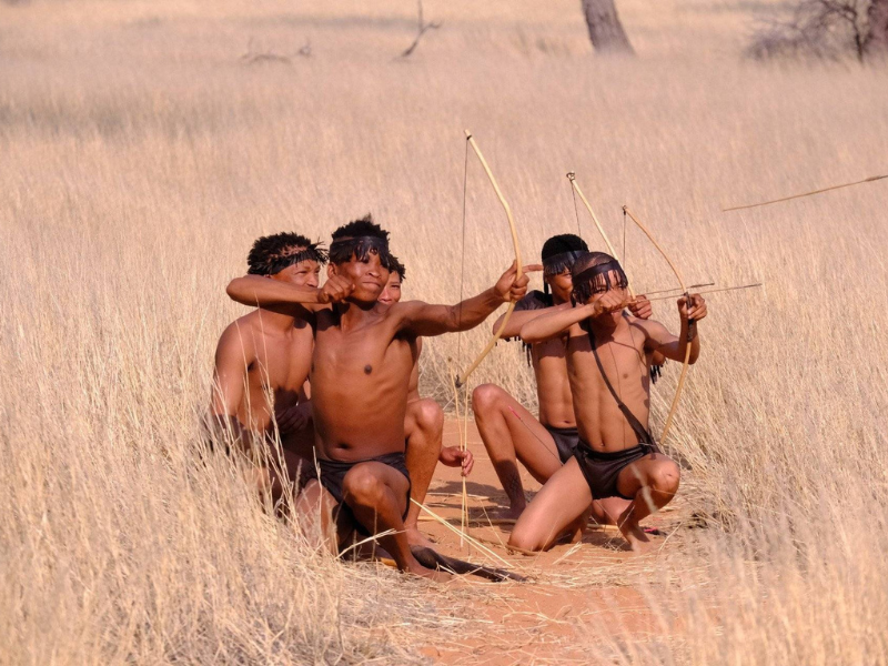 San Bushmen experience demonstrating traditional bow-and-arrow hunting techniques in a dry grassy landscape.
