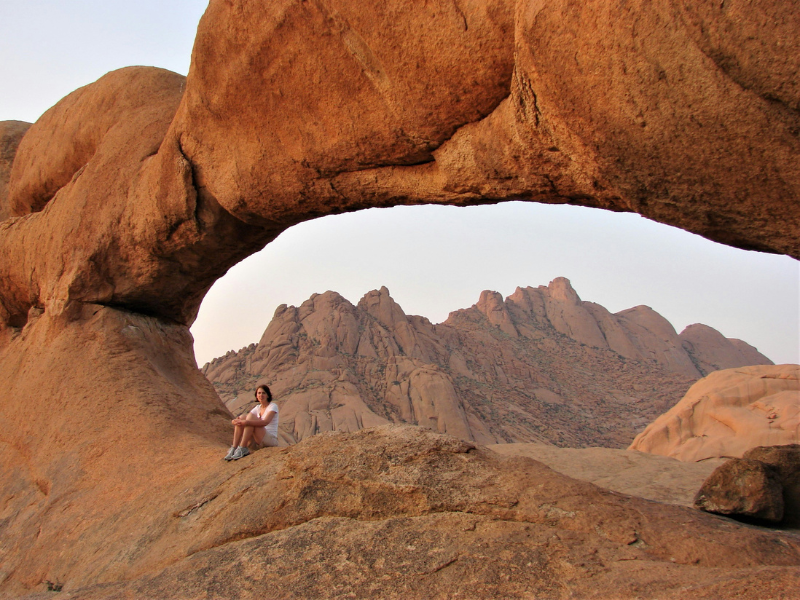 A person sitting beneath a natural rock arch at Spitzkoppe with jagged mountains in the background.