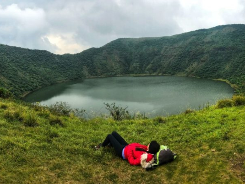 Hikers walking along a lush green mountain trail with Lake Burera and rolling hills in the background.