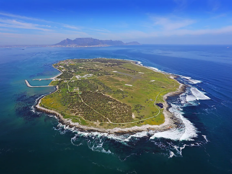 Aerial view of Robben Island surrounded by blue ocean with Cape Town and Table Mountain visible in the distance.