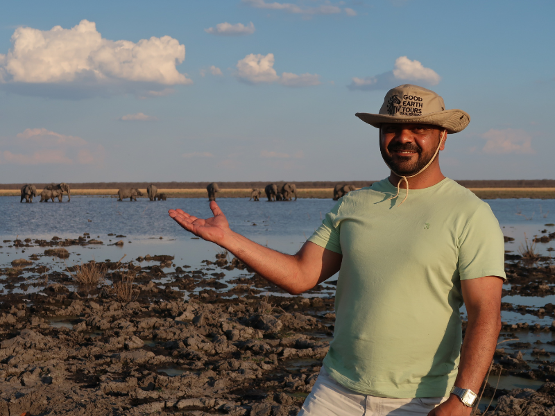 Good Earth Tours guide smiling by a floodplain, gesturing toward elephants in the distance