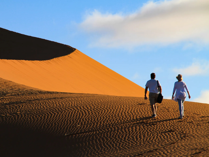Two travelers walking along a towering Namibia sand dune under a bright blue sky