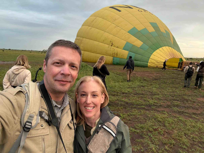Couple on safari taking a selfie in front of a hot air balloon being prepared at sunrise in the savannah, capturing the excitement of an early morning adventure and planning how far in advance to book African safari experiences.