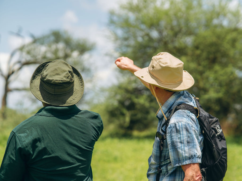 Safari guide and traveler wearing hats and backpacks, pointing out wildlife in the distance during a guided walking safari experience in the African bush.