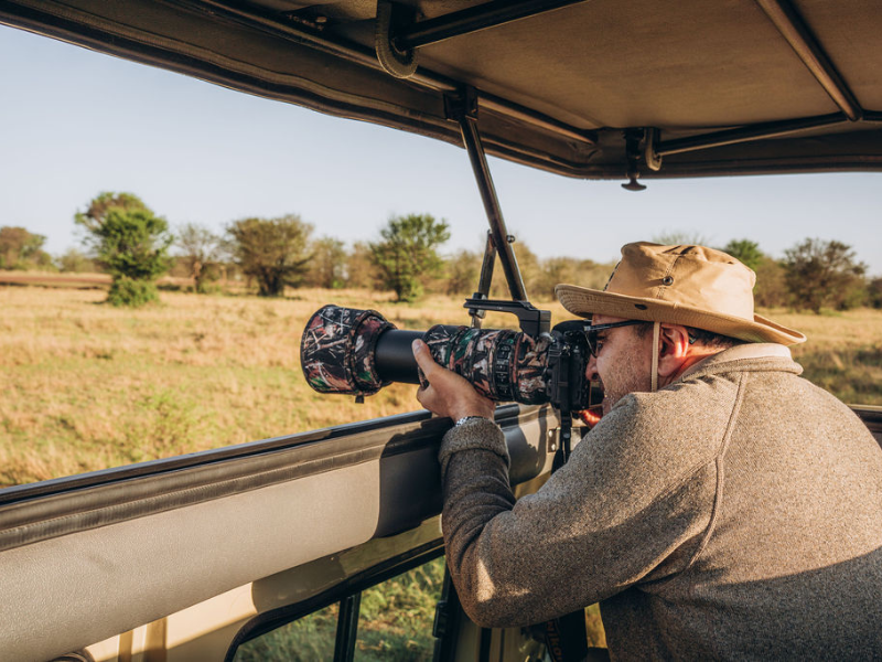 Photographer in an open safari vehicle using a telephoto lens to capture wildlife, demonstrating the importance of having the right gear for game drives.