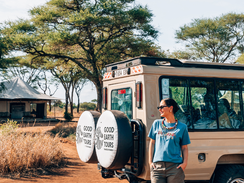 Traveler standing beside a safari vehicle at camp, enjoying a relaxed moment in nature far from typical resort settings.