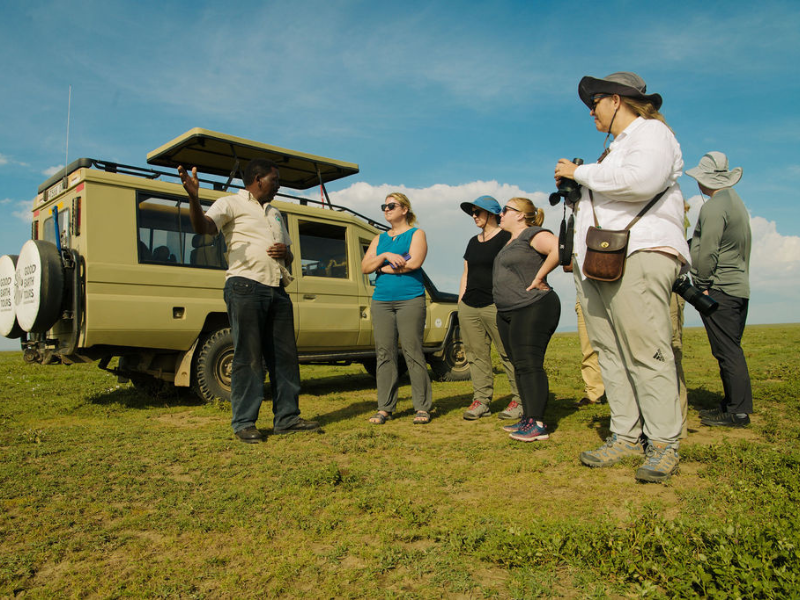Safari guide briefing a group of travelers beside a vehicle in open plains, answering common questions like do I need a safari planner before heading out on a game drive.