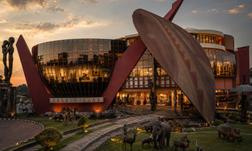 Striking cultural heritage centre building at dusk, its curved glass facade and dramatic red and horn-shaped roof structure glowing with interior lights, surrounded by bronze wildlife sculptures and a reflecting pool.