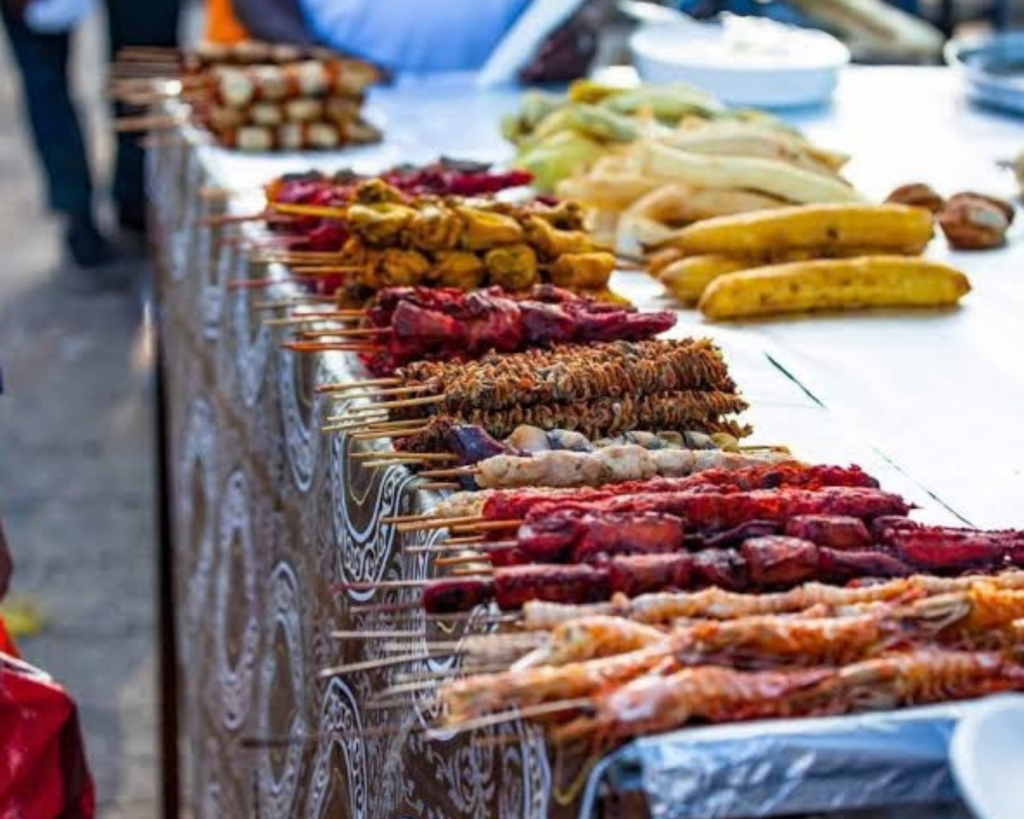 Rows of grilled skewers—meat, chicken, seafood, and shrimp—laid out alongside roasted plantains and corn on a decorative tablecloth.