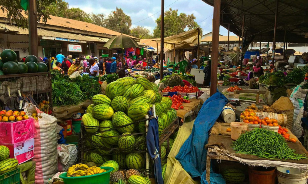 Bustling produce section of the market with towering piles of watermelons, bananas, avocados, tomatoes, and green beans displayed under a corrugated metal roof, shoppers gathered in the background.