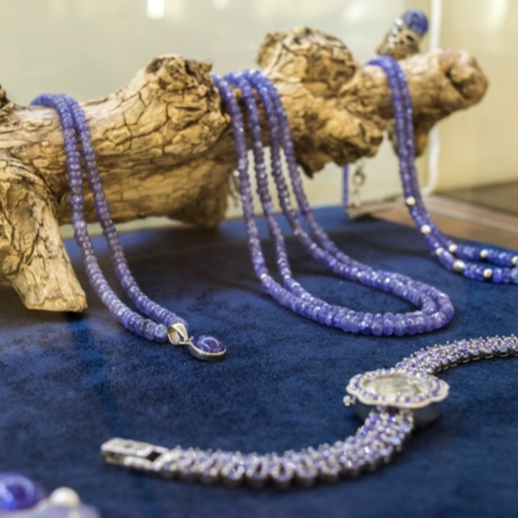 Multiple strands of tanzanite beaded necklaces draped over a driftwood display stand at the Tanzanite Experience museum, alongside a pendant and an ornate beaded bracelet on blue velvet.